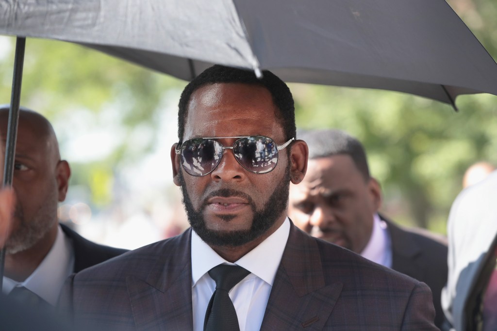 R. Kelly in a suit standing under an umbrella with sunglasses on 